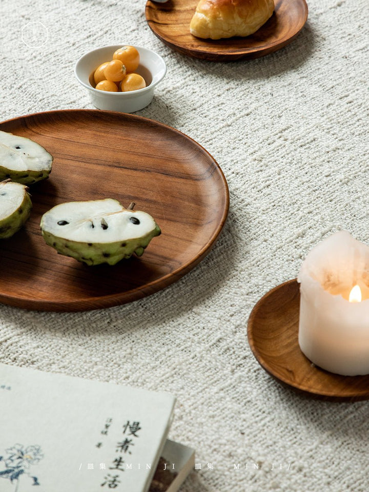 Handcrafted teak wood plates with fruit and rolls on textured fabric.