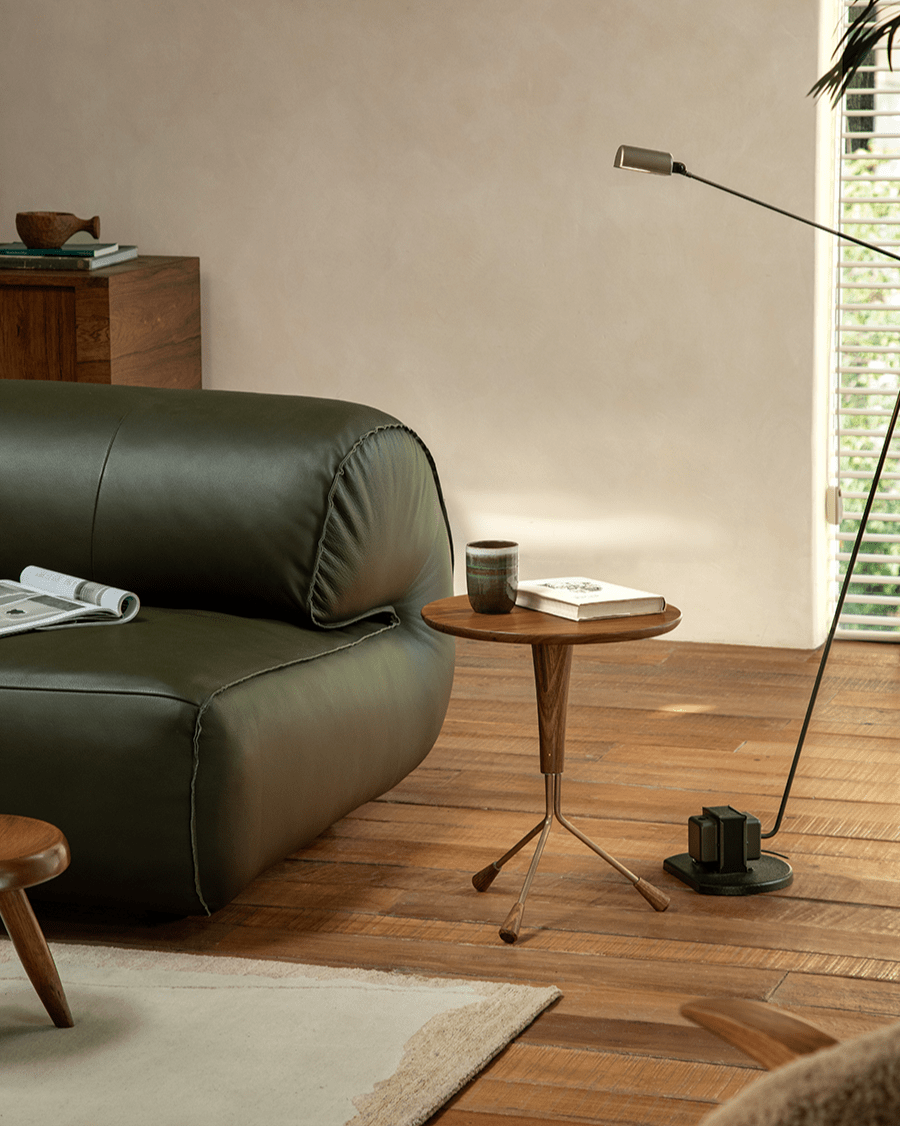 Larsson Side Table with round wooden top and tripod base, next to a dark green leather sofa
