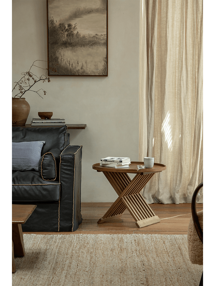 Round teak folding coffee table with slatted design beside a black sofa in a bright living room.
