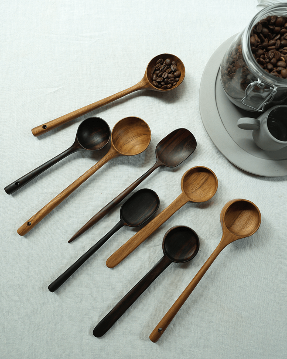 Teak coffee spoons in varying shades of brown and black on a white linen surface, with coffee beans.