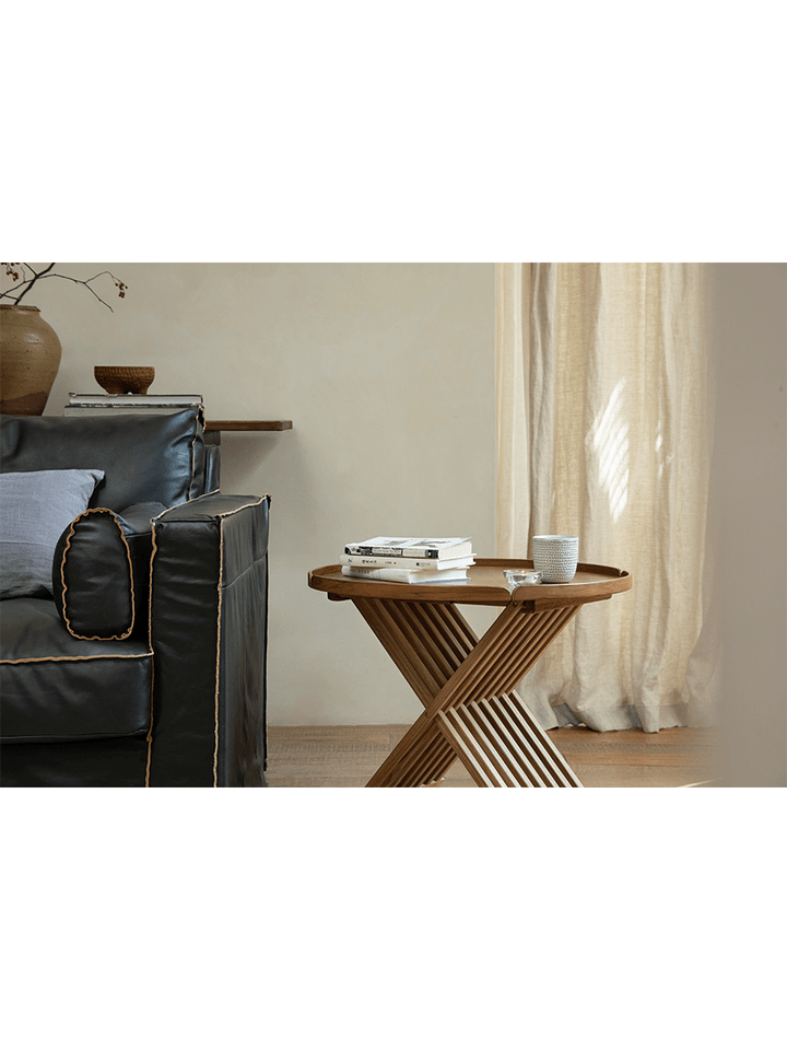 Round teak folding coffee table with slatted base next to a black sofa in a bright living room