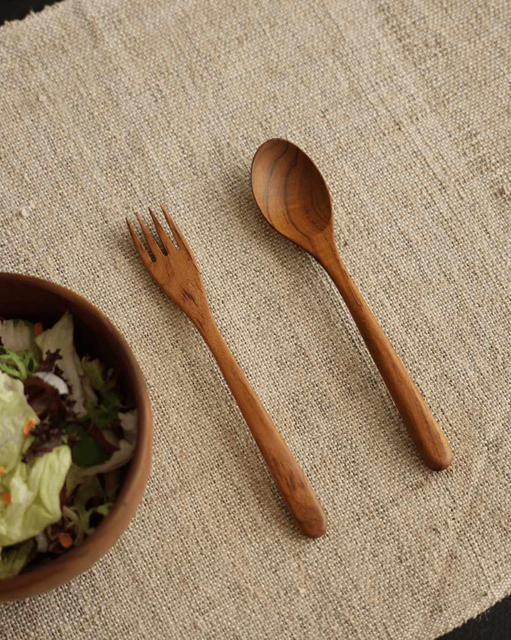 Wooden spoon and fork set with salad bowl on linen fabric, natural wood finish, product shot