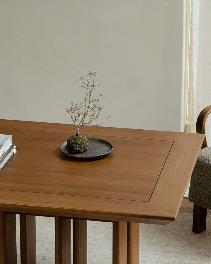 Minimalist teak dining table with small plant and books in a softly lit interior.