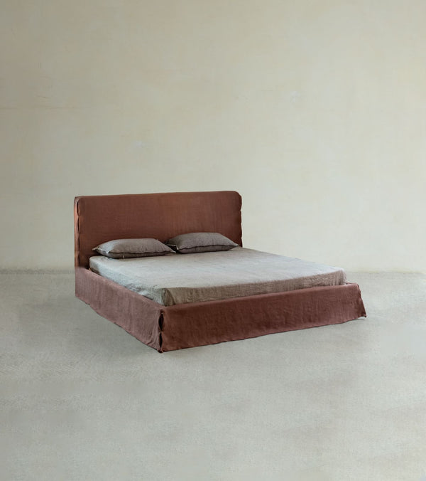 The Freya Linen Bed by ZM Home features a minimalist solid pine frame, a brown upholstered headboard, and two pillows, set against a plain beige wall and floor.