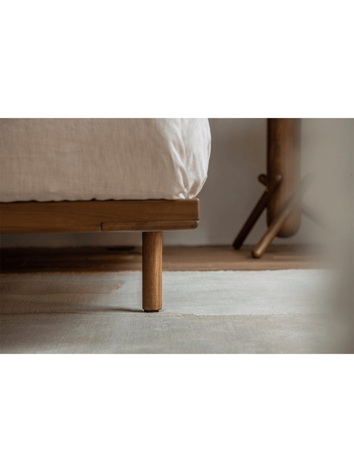 Teak bed frame leg detail on textured rug in minimalist bedroom setting.