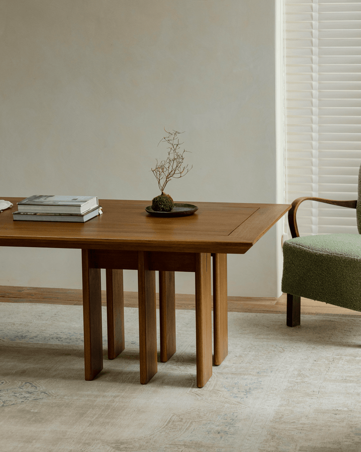 Titan teak dining table with multi-leg design, decorative plant, and books in bright interior.