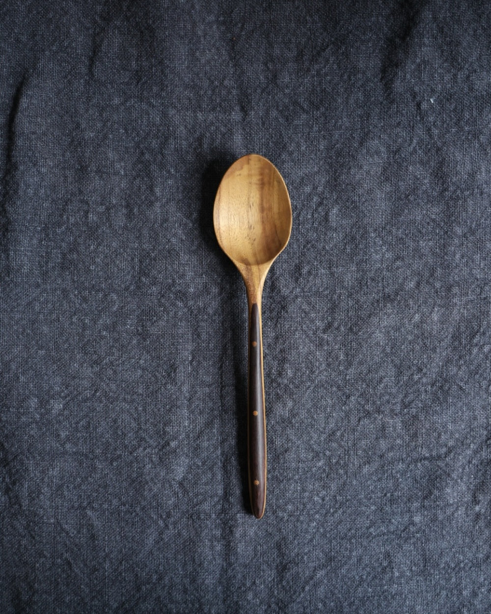 Ebony wood handle table spoon with teak spoon head and dotted detail on dark fabric.