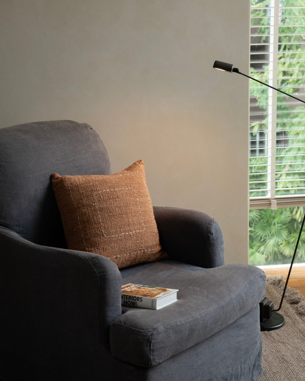 Handwoven Sylia Pillow in rust orange on grey armchair with "Interiors Now!" book