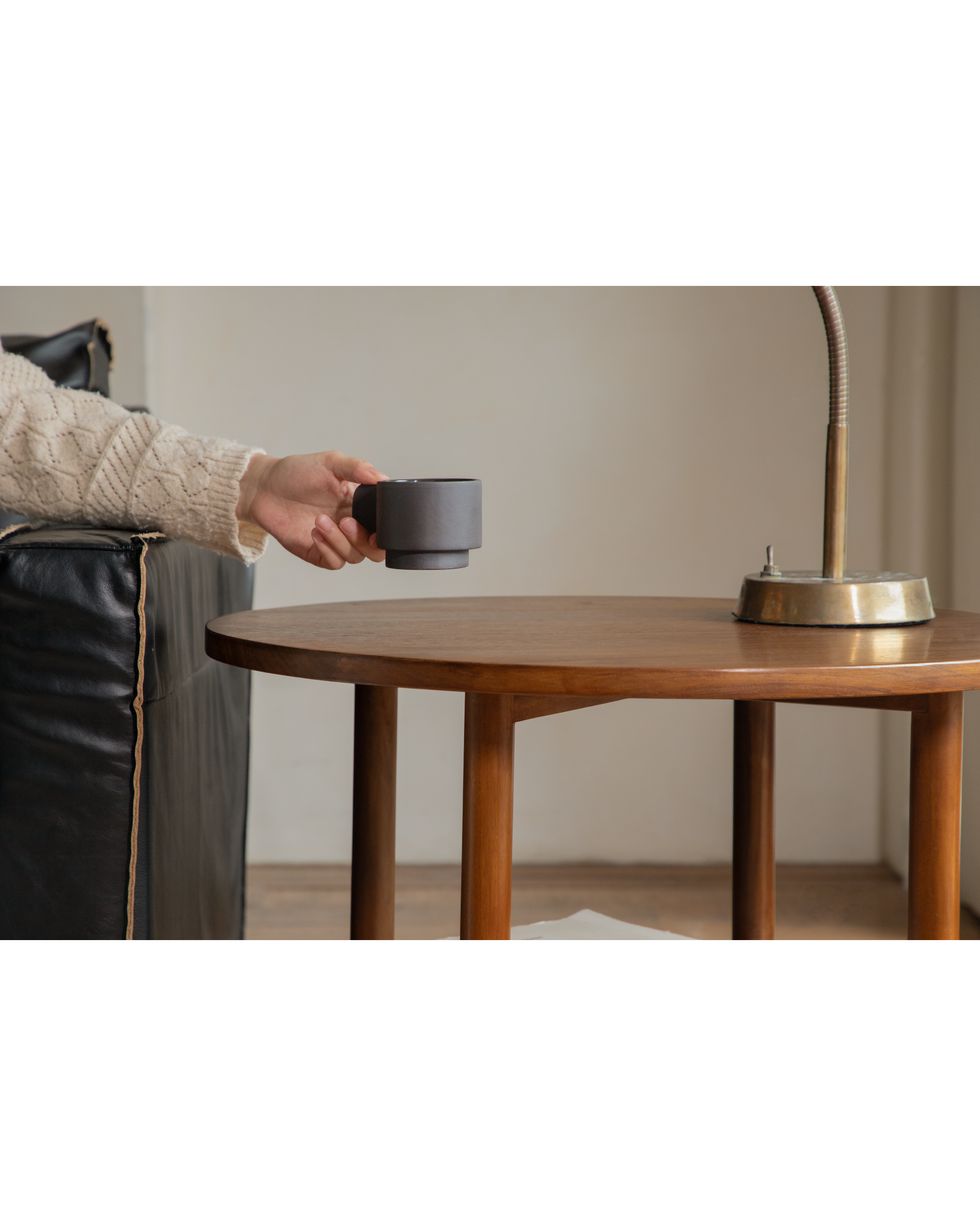 Daisy side table, round teak wood, with brass lamp and hand holding mug.