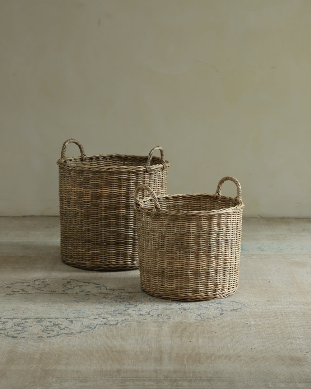 Two Aria round woven rattan storage baskets with handles on a patterned rug.