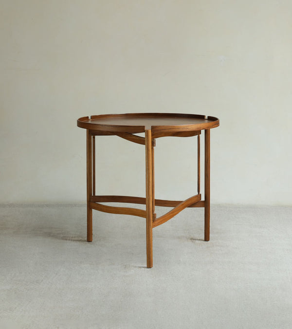 The Hansen Tray Side Table by ZM Home, featuring a round wooden top with raised edge and sturdy triangular base, stands on a light carpet against a plain off-white wall.