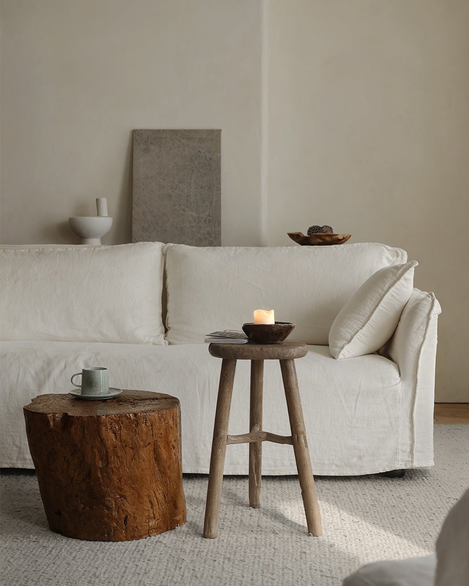 A minimalist living room featuring a white sofa, a stump coffee table with a mug, and the ZM Home Reclaimed Wood Vintage Chinese Stool holding a candle, all set against neutral-toned decor and a light wall.