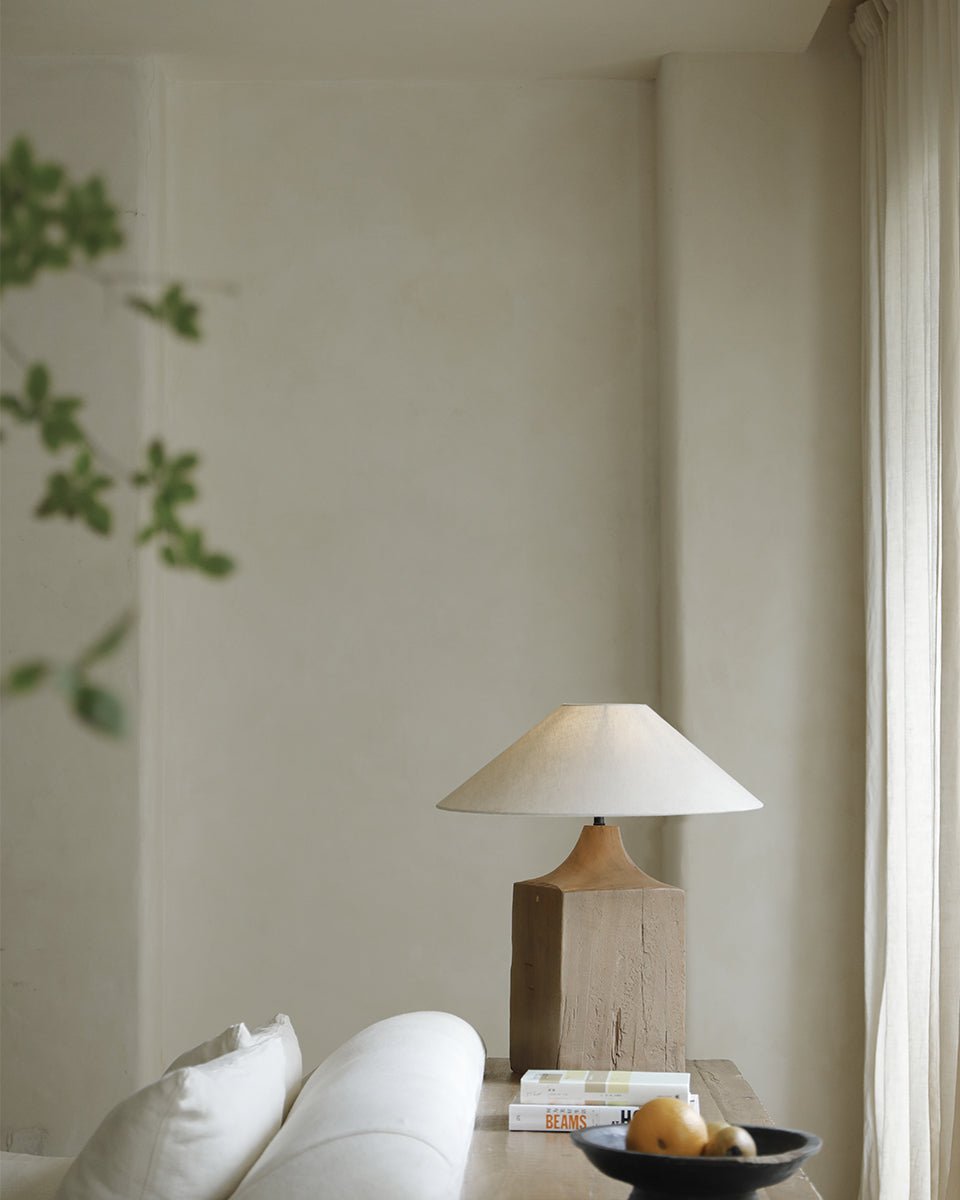 Wooden table lamp with beige linen shade, on wooden table with books and fruit bowl, cozy living room