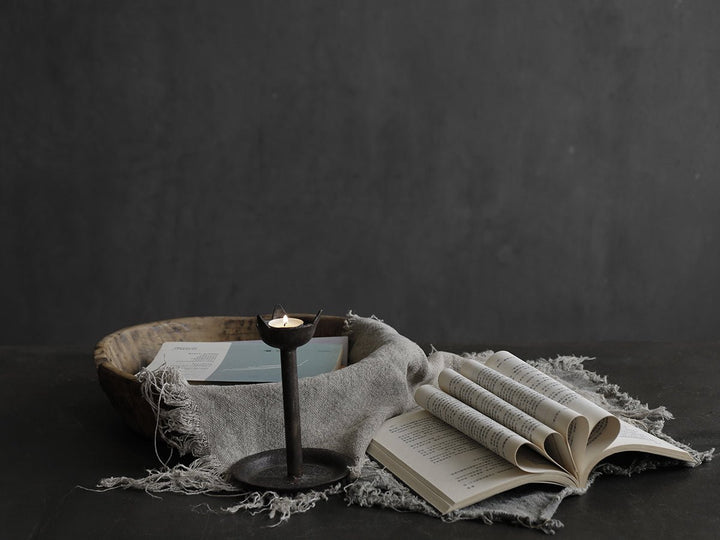 Rustic iron candle holder with soft textiles and open books in a cozy setting.