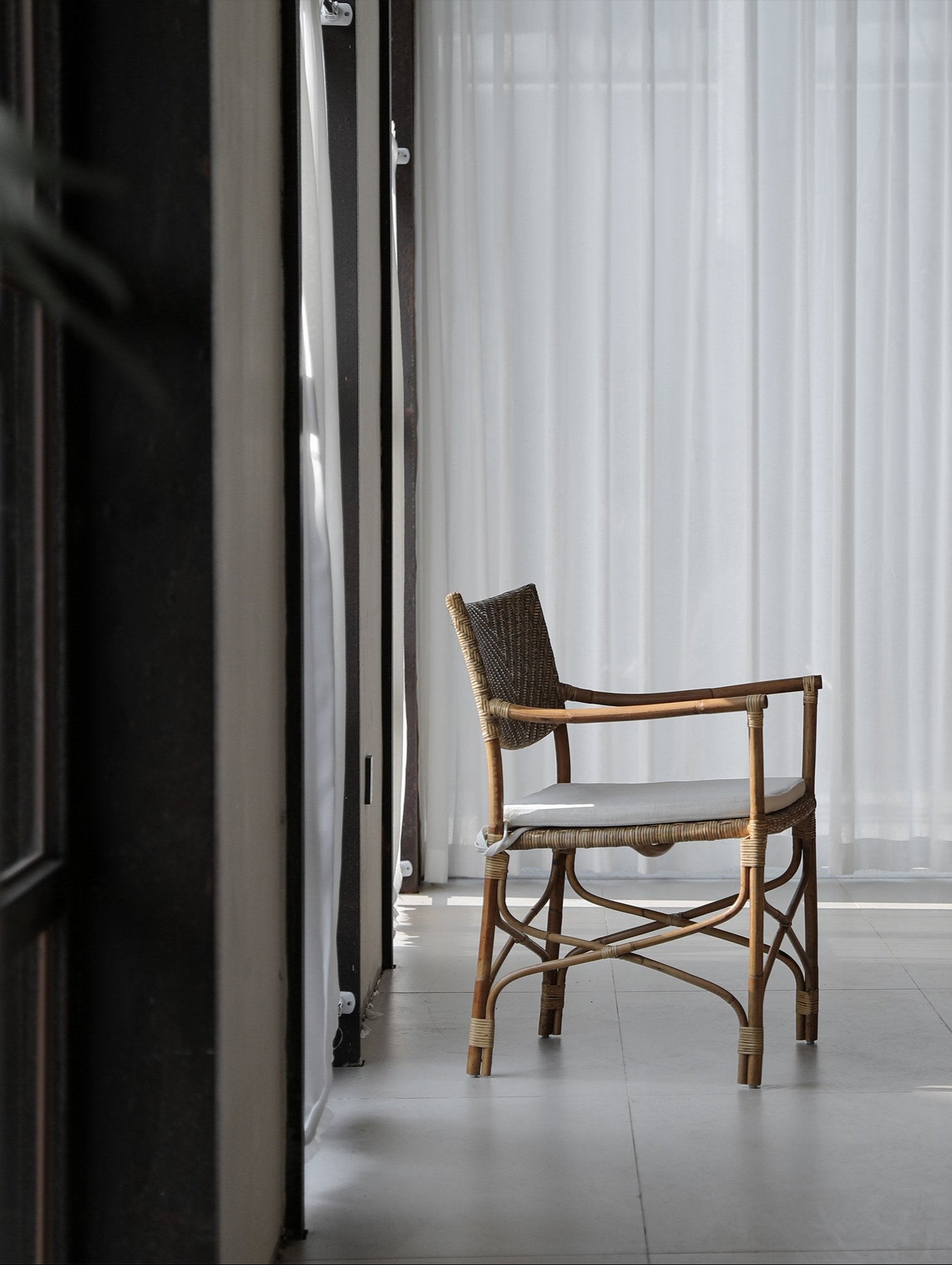 Dingee Chair with woven back, natural wood frame, and cushion, next to sheer white curtains in a bright room