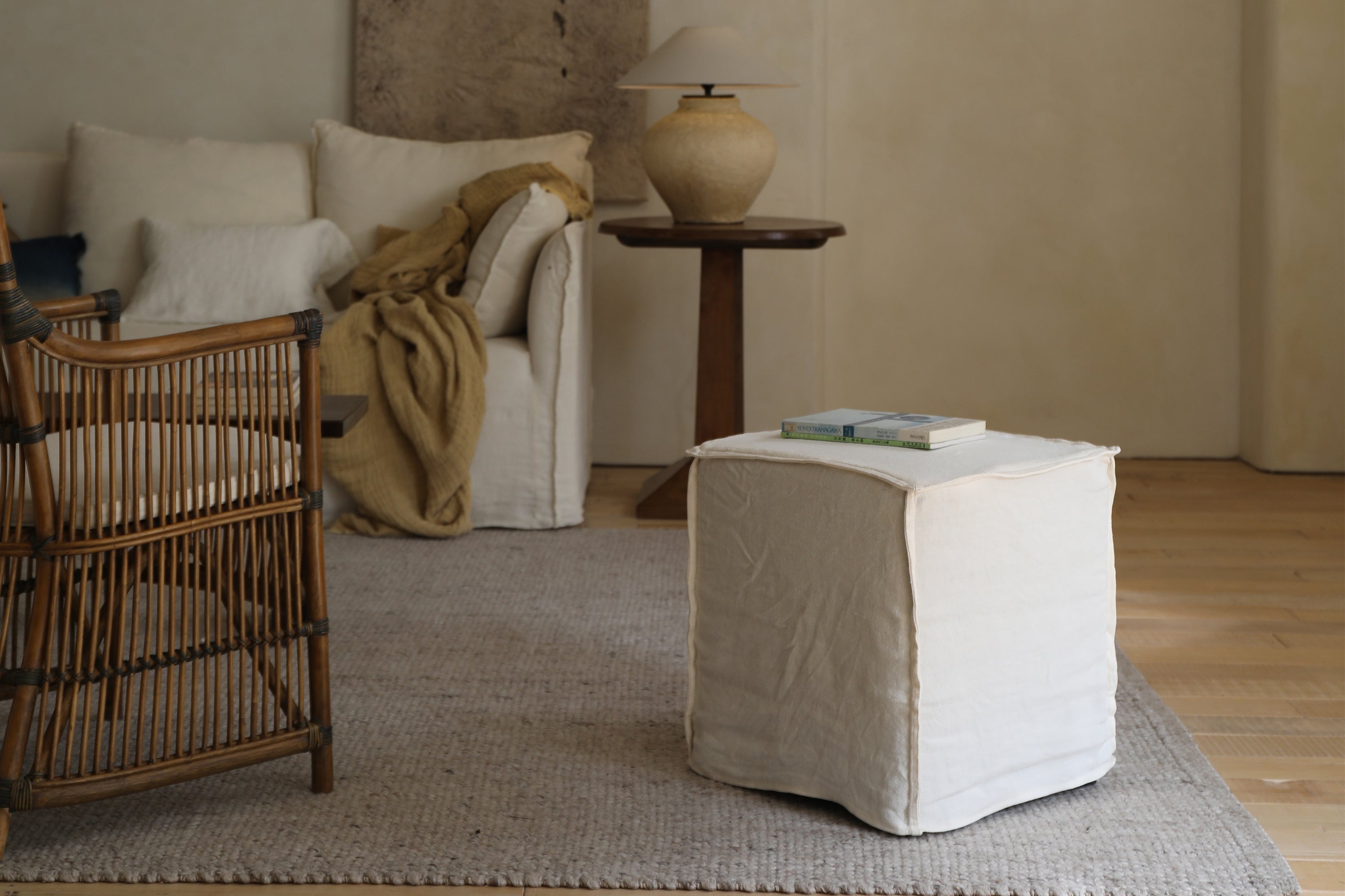 A ZM Home Cube Linen Ottoman with two books on top rests on a textured rug in a softly lit room featuring wooden flooring.
