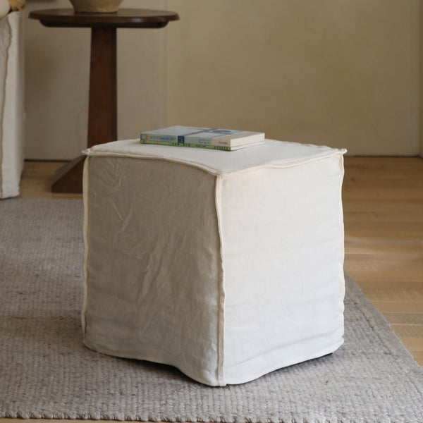 A ZM Home Cube Linen Ottoman with two books on top rests on a textured rug in a softly lit room featuring wooden flooring.