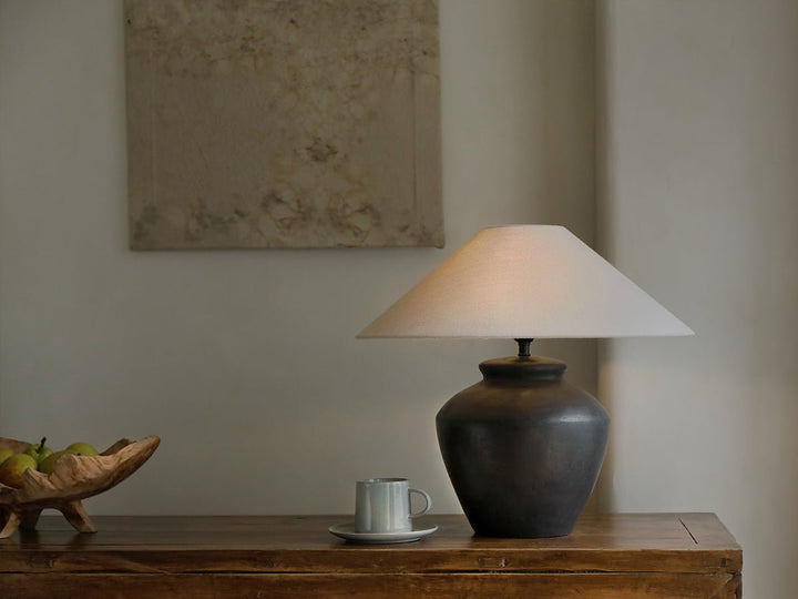 Dark vase lamp with light shade on wooden table beside cup and fruit bowl.